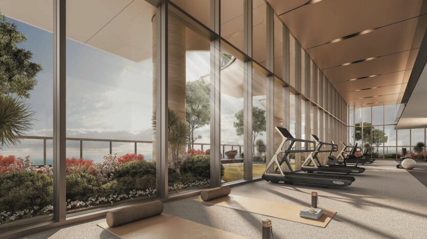 Une salle de sport moderne du SkyParks avec de grandes fenêtres allant du sol au plafond, des tapis d'exercice, des bouteilles d'eau et des tapis roulants. Des fleurs et de la verdure luxuriante sont visibles à l'extérieur, offrant une vue pittoresque et la lumière naturelle du soleil.