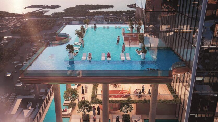 Une piscine à débordement sur le toit, avec des chaises longues et des personnes se relaxant aux SkyParks, avec vue sur un paysage urbain, l'eau et des îles lointaines. Les parois vitrées de la piscine couronnent un immeuble moderne de grande hauteur avec une terrasse en contrebas.