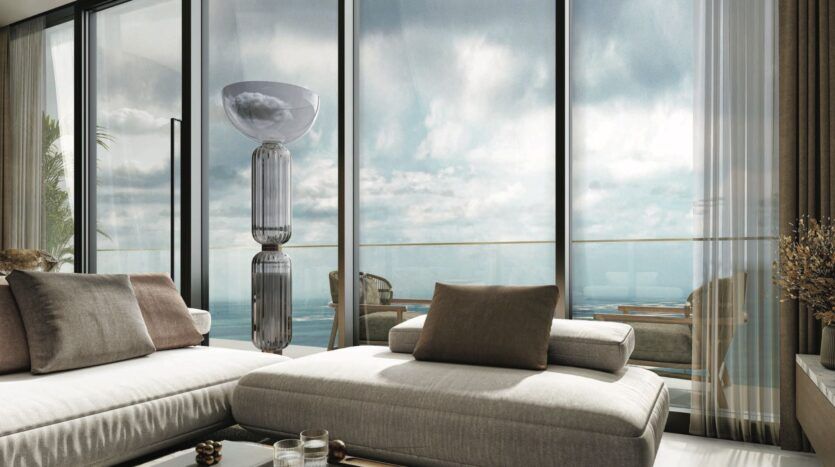 Salon moderne avec des canapés beiges et des coussins neutres, de grandes fenêtres allant du sol au plafond, une lampe haute et une vue sur un balcon extérieur du SkyParks donnant sur l'océan sous un ciel nuageux.