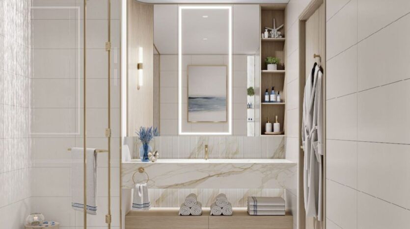 Salle de bains moderne à la Résidence Sunset Bay Grand par Imtiaz à Dubai Islands avec un grand miroir éclairé, un comptoir en marbre, des étagères ouvertes avec des serviettes et des articles de toilette, une douche en verre et des carreaux blancs. Un peignoir est accroché au mur, reflété par des œuvres d'art encadrées.