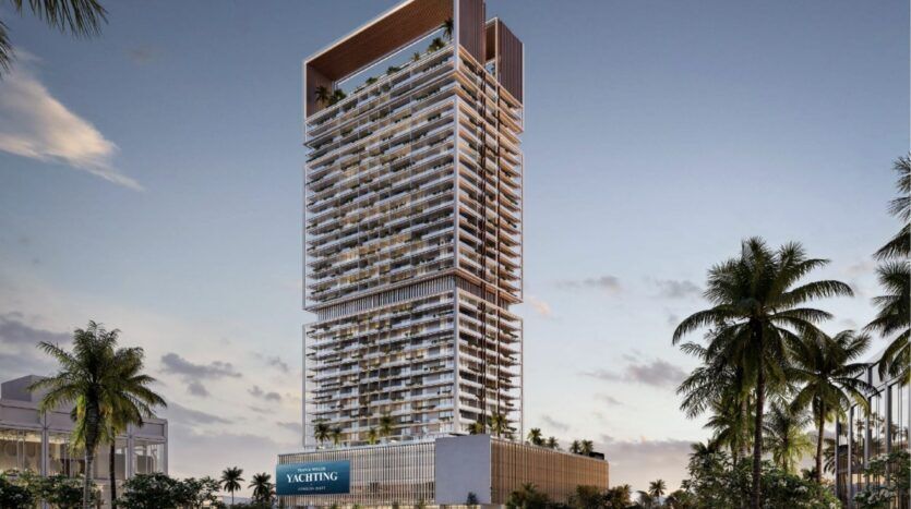 Une tour moderne dotée de nombreux balcons et d'une terrasse sur le toit se dresse au milieu des palmiers au coucher du soleil dans la ville maritime de Dubaï. La partie inférieure comporte un panneau "YACHTING", évoquant l'élégance de Franck Muller Yachting à Dubai Maritime City. Le ciel est partiellement nuageux.