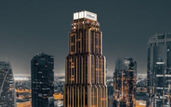 Grand gratte-ciel moderne surmonté de l'enseigne "Hilton", les résidences Hilton se détachent sur un paysage urbain de gratte-ciel à la tombée de la nuit, avec des accents dorés et sombres saisissants.