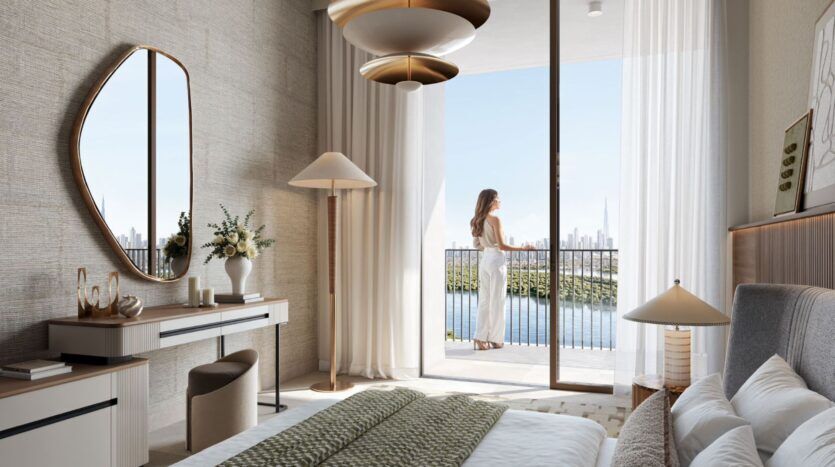 Modern bedroom with neutral tones, a large bed, and a desk with a mirror. A woman stands on the outdoor balcony, which overlooks Creek Bay and the city skyline through floor-to-ceiling glass doors. Natural light fills this elegant space.