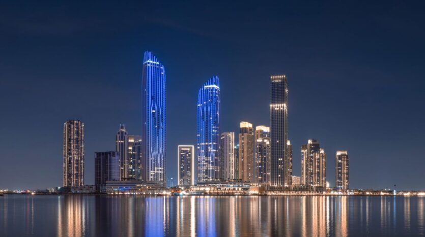 The skyline of a modern city at night, with tall illuminated skyscrapers and blue and white lights reflecting on the calm waters of Creek Bay in the foreground. The sky is dark and clear.