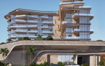 Un bâtiment moderne de plusieurs étages avec des balcons en verre incurvés, des murs texturés de couleur beige et des terrasses verdoyantes vous accueille à Flora Bay. Des palmiers et des voitures garées bordent l'entrée sous un ciel bleu clair.