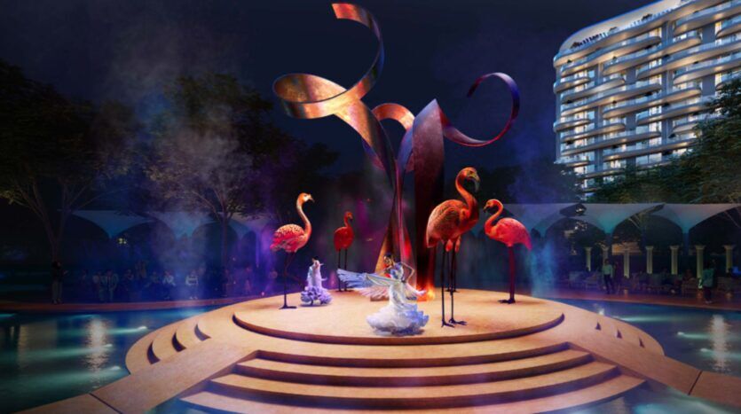 Una escena nocturna en la Piazza Roma muestra a bailarines vestidos con trajes blancos en un escenario circular rodeado de grandes esculturas de flamencos rosados, con una pieza central abstracta curvada y un edificio iluminado al fondo.