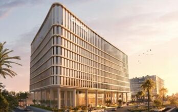 Un immeuble de bureaux moderne à plusieurs étages avec des façades en verre se dresse au coucher du soleil près de la Piazza Roma, entouré de palmiers et d'allées paysagères. Des gens se promènent à l'extérieur tandis que des oiseaux volent au-dessus de leur tête. Un autre bâtiment similaire est visible à l'arrière-plan.