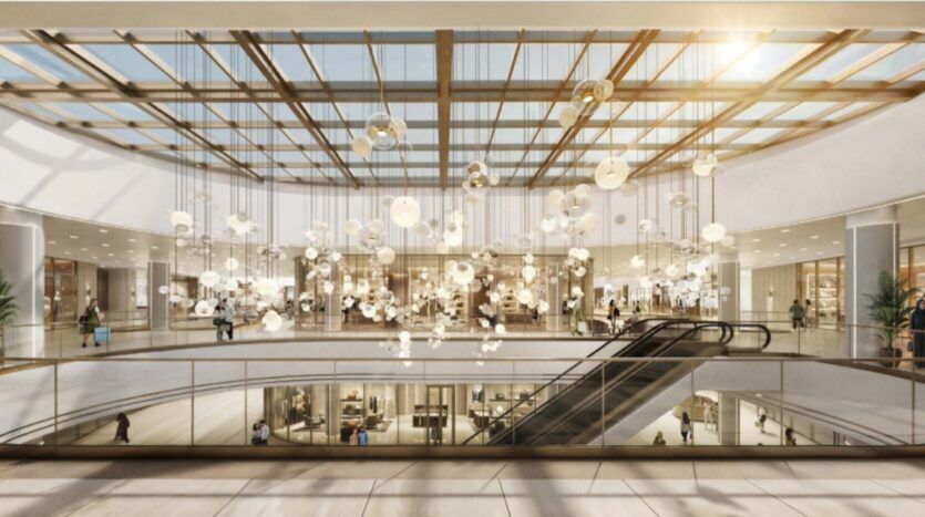 El interior de un moderno centro comercial inspirado en la Piazza Roma, con un gran techo de cristal, luces colgantes, escaleras mecánicas y tiendas en dos niveles. La luz del sol entra por las ventanas, creando un ambiente luminoso y acogedor para los visitantes.