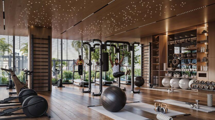 Moderno gimnasio con suelos de madera, aparatos de ejercicio dispersos, balones medicinales y dos personas entrenando. Grandes ventanales con vistas a la Piazza Roma y a las palmeras. El techo está iluminado por pequeñas luces en forma de estrellas que resaltan la luz natural del exterior.