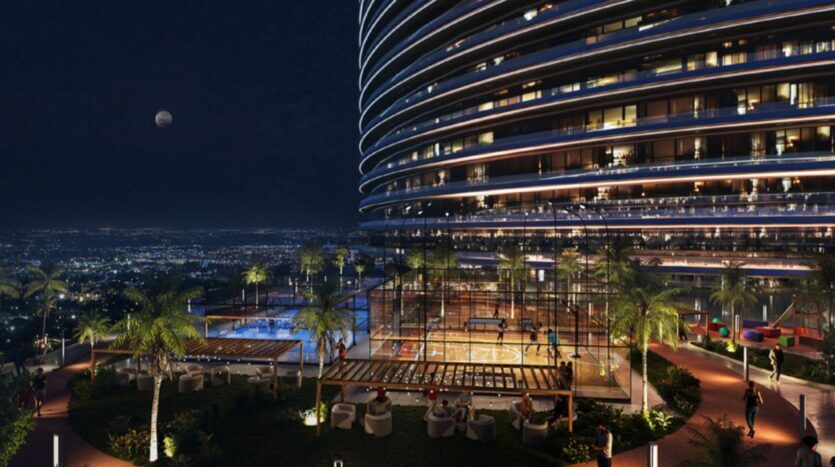 Une tour moderne illuminée la nuit surplombe un paysage urbain. La terrasse, connue sous le nom de Skyflame, comprend des palmiers, un terrain de basket, un court de tennis et des places assises en plein air, le tout sous un éclairage doux et avec la lune visible dans le ciel.