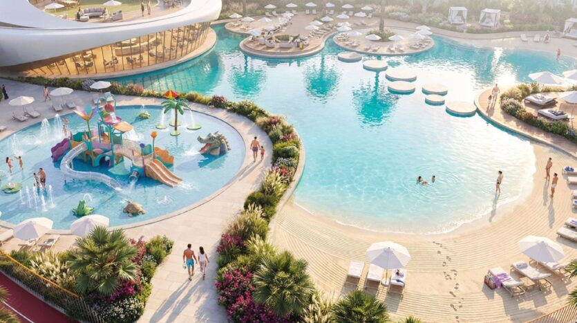 Vue aérienne de la piscine du complexe Greenz by Danube, avec une plage de sable à l'entrée, des chaises longues, des parasols et une aire de jeux colorée pour les enfants. Plusieurs personnes se baignent et se détendent, entourées de plantes et de jardins luxuriants.