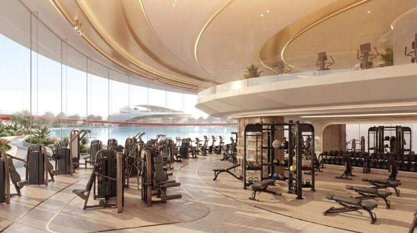 Salle de sport spacieuse et moderne à Greenz by Danube avec de grandes fenêtres, une lumière naturelle abondante, diverses machines d'exercice, des poids libres, et une disposition ouverte et incurvée avec un niveau supérieur. À l'extérieur, on aperçoit un bâtiment futuriste.