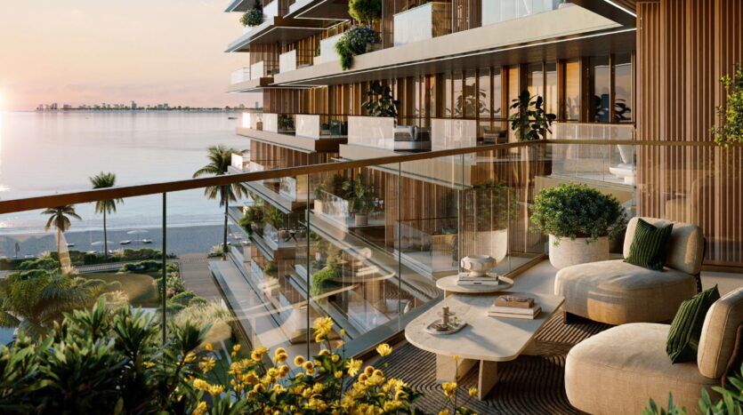 Balcon luxueux des Résidences Cheval avec mobilier moderne, balustrade en verre, plantes en pot et table basse, donnant sur une plage sereine et l'océan au coucher du soleil. Les fleurs et les palmiers rehaussent l'atmosphère élégante et relaxante.