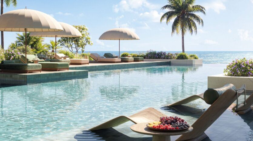 Piscine à débordement surplombant l'océan aux Résidences Cheval, avec chaises longues, parasols et plateau de fruits frais. Une végétation luxuriante et des palmiers entourent la piscine, créant une atmosphère tropicale relaxante sous un ciel clair et lumineux.