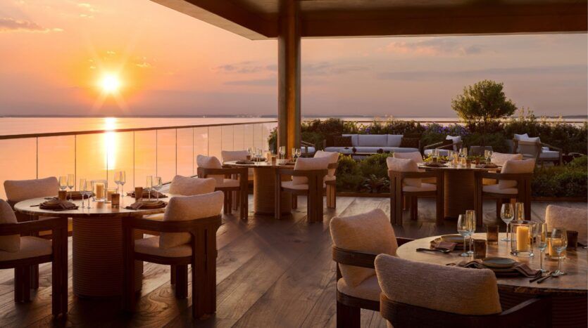 Élégante terrasse du restaurant extérieur des résidences Cheval, surplombant un plan d'eau calme au coucher du soleil, avec des tables dressées, des chaises rembourrées, une balustrade en verre et un éclairage doux et chaleureux créant une atmosphère sereine et invitante.