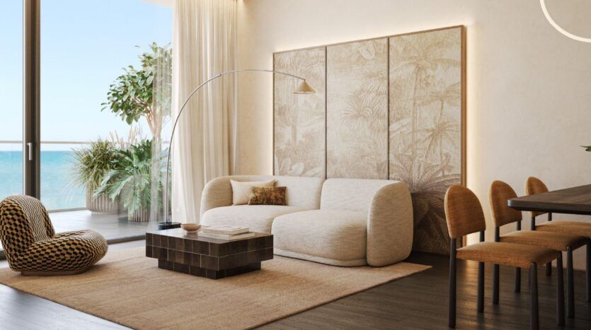 Le salon moderne de Cheval Residences est composé d'un canapé crème, d'une chaise longue à motifs et d'une table basse en bois foncé. De grandes fenêtres offrent une vue sur l'océan, tandis que les tons neutres et l'art mural créent une atmosphère calme et aérée.