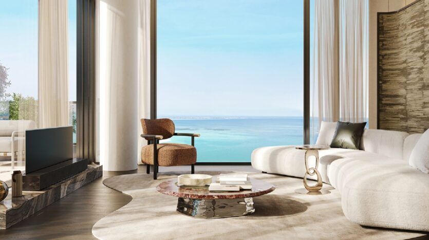 Un salon moderne aux Résidences Cheval avec de grandes fenêtres offrant une vue panoramique sur l'océan. L'espace est composé d'un canapé blanc incurvé, d'un fauteuil marron, d'une table basse en marbre et d'un décor neutre où la lumière naturelle pénètre à flots.