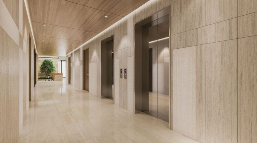 Un couloir moderne à Derby Heights présente des murs en bois clair, des sols en carrelage poli et trois portes d'ascenseur en acier inoxydable. Un arbre vert en pot ajoute une touche de nature au bout du couloir.