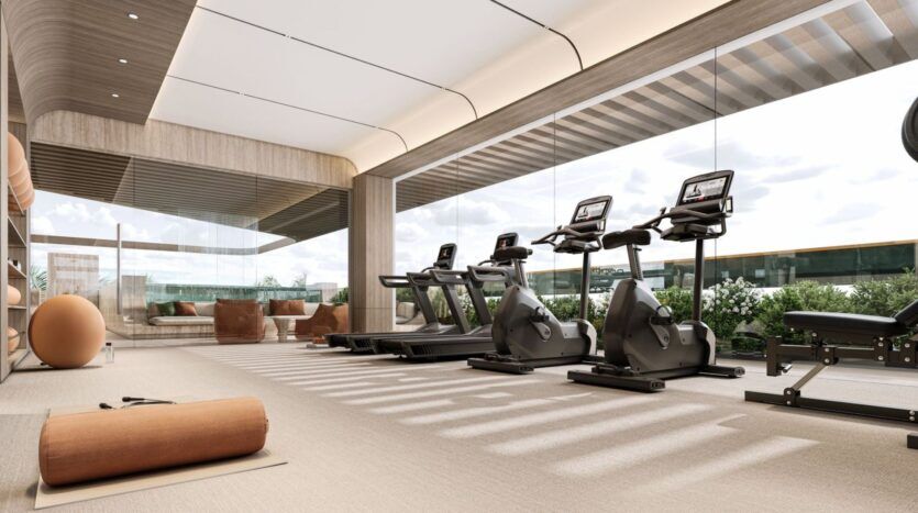 La salle de sport moderne de Derby Heights, dotée de grandes fenêtres, est équipée de tapis de course, de vélos d'appartement, de ballons d'exercice, de tapis de yoga et d'un espace pour s'asseoir. L'espace est baigné de lumière naturelle et la verdure est visible à l'extérieur.