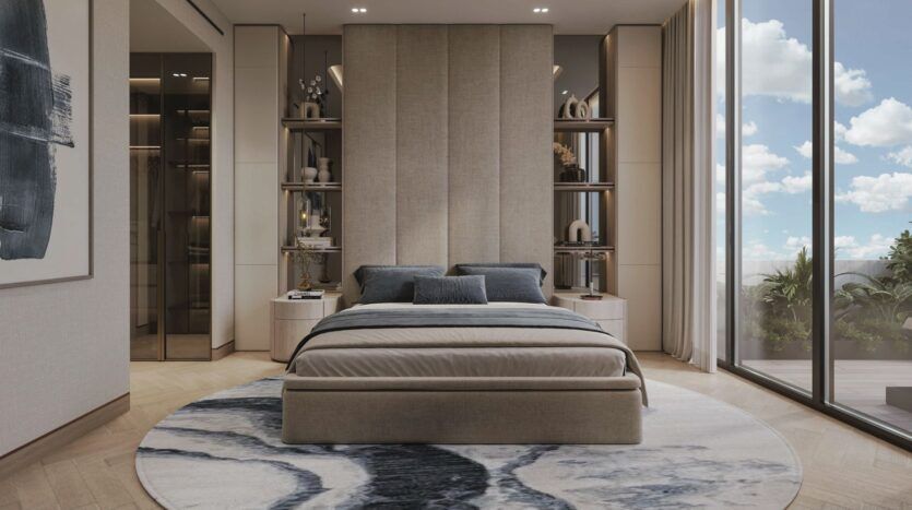 Chambre à coucher moderne aux Résidences Meriden Beach avec un grand lit rembourré, une literie grise et beige, des étagères encastrées, une fenêtre allant du sol au plafond et montrant un ciel bleu, et un tapis à motifs ronds sur un sol en bois. La décoration est minimaliste et dans des tons neutres.