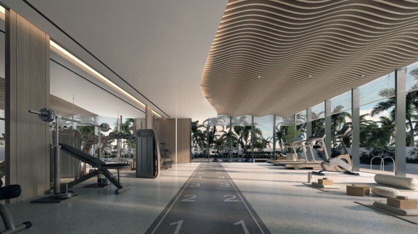 La salle de sport moderne des résidences Meriden Beach est dotée de grandes fenêtres donnant sur les palmiers. Elle comprend des machines d'exercice, des poids libres, des tapis de course, des tapis de yoga et une piste d'athlétisme. Le plafond ondulé et la lumière naturelle créent une atmosphère accueillante.