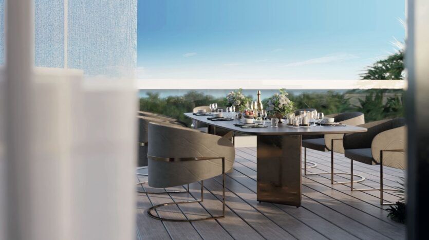 Salle à manger moderne en plein air sur une terrasse en bois des résidences Meriden Beach, avec une table rectangulaire et des chaises grises élégantes, donnant sur une végétation luxuriante et un ciel bleu limpide.
