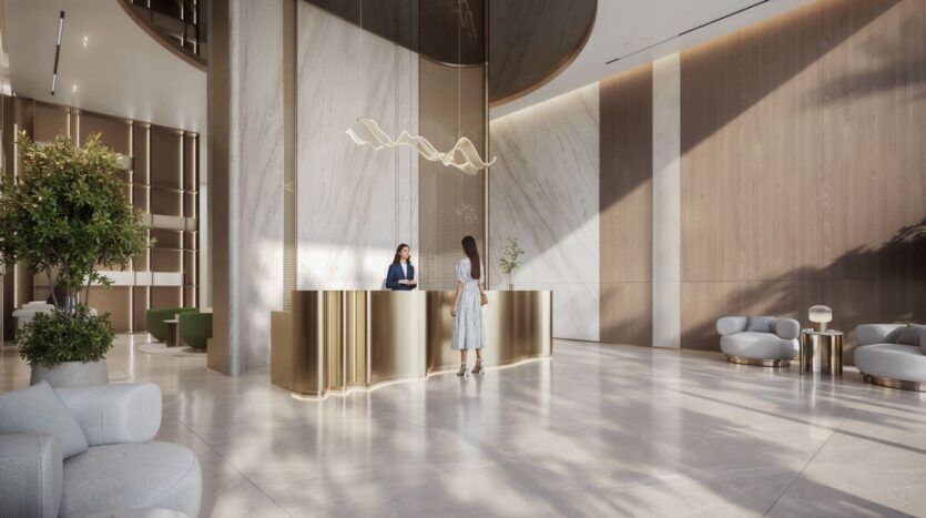 Un hall d'hôtel moderne avec des sols en marbre, un comptoir de réception en or et un éclairage élégant accueille les clients de The Tides by Amis. Deux femmes sont à la réception, entourées de plantes luxuriantes et de sièges élégants dans un espace lumineux et spacieux.