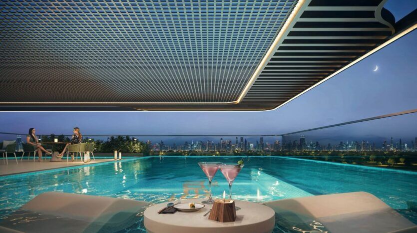 Une luxueuse piscine sur le toit au crépuscule au Tides by Amis, avec vue sur la ville, deux cocktails roses et un dessert sur une table dans l'eau. Deux personnes sont assises au bord de la piscine alors qu'un croissant de lune brille dans le ciel clair du soir.
