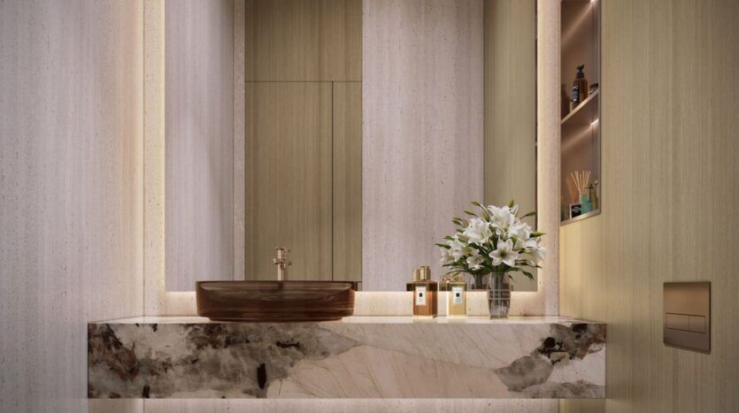Salle de bains moderne avec un comptoir en marbre, un évier en verre brun et un robinet en or. Un exemplaire de The Tides d'Amis est posé à côté d'un vase de lys blancs, de flacons de parfum et de produits de toilette, sous un éclairage d'ambiance doux.