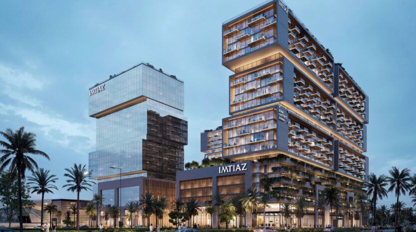 Raw District by Imtiaz est un complexe immobilier moderne composé de deux tours principales avec des façades en verre et de grandes terrasses, entourées de palmiers. Le nom "IMTIAZ" est affiché sur les deux tours. La scène du crépuscule est éclairée par des lumières intérieures accueillantes.