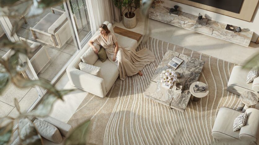 Une femme vêtue d'une longue robe beige est assise sur un canapé crème dans un salon moderne Tilal by Binghatti, doté de grandes fenêtres, de tables en marbre et d'un tapis à motifs, vu d'en haut à travers des branches feuillues.