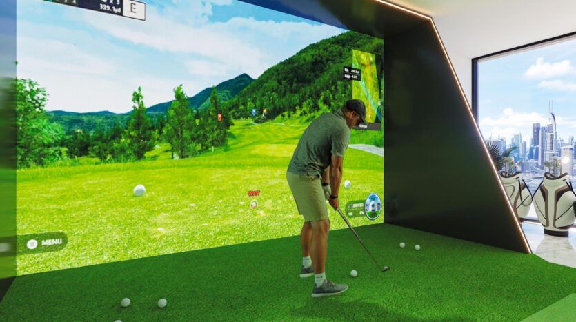 Une personne joue au golf sur un simulateur de golf en salle équipé d'un grand écran affichant un parcours de golf virtuel, au Vue Dorée. Plusieurs balles de golf sont posées sur le gazon vert, et on aperçoit des sacs de golf et des clubs près d'une fenêtre donnant sur la ville.
