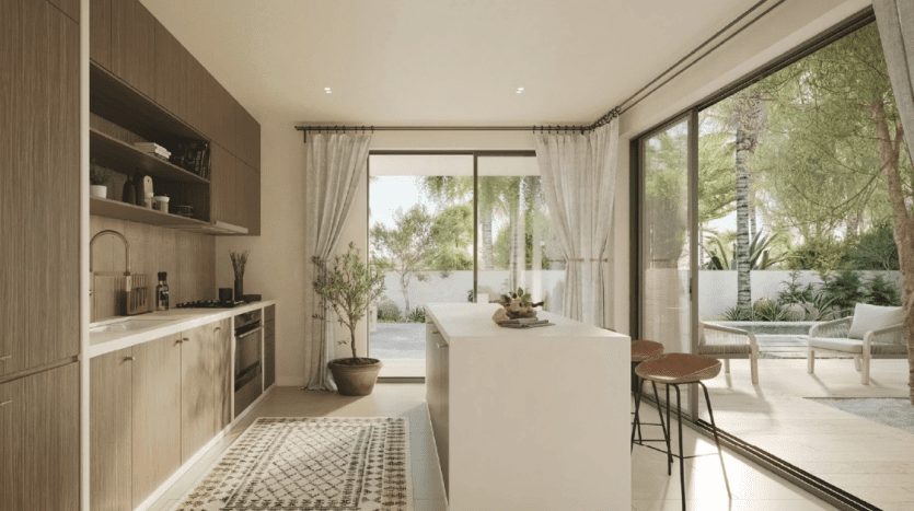 Modern kitchen interior with elegant wood cabinets and an island, leading to a sunny patio overlooking outdoor green plants and a sitting area. A floor-to-ceiling transparent door enhances the natural light, epitomizes
