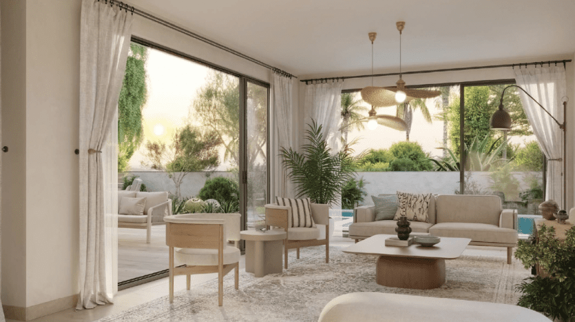 A modern living room in a Dubai villa with large glass doors opening onto a garden, featuring neutral-toned furniture, a large rug and a ceiling fan, bathed in warm light.