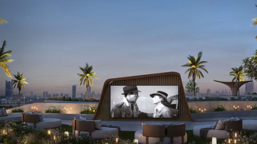 Un cinéma en plein air sur le toit, au coucher du soleil, aux Mövenpick Residences : un grand écran diffuse un film en noir et blanc, entouré de sièges confortables, de palmiers, avec la silhouette de la ville en arrière-plan. De petites lumières créent une ambiance chaleureuse et accueillante.