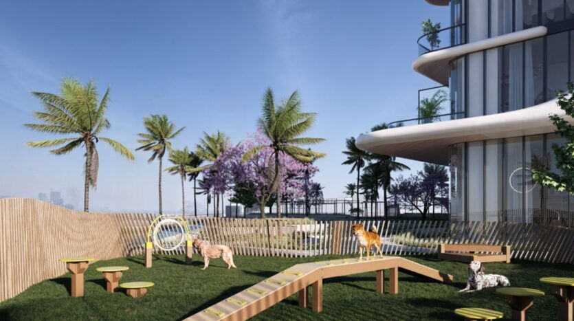 Un parc canin en plein air moderne situé au sein des Mövenpick Residences comprend des structures de jeux en bois, trois chiens, des palmiers et, en arrière-plan, un bâtiment aux vitres incurvées sous un ciel bleu limpide.