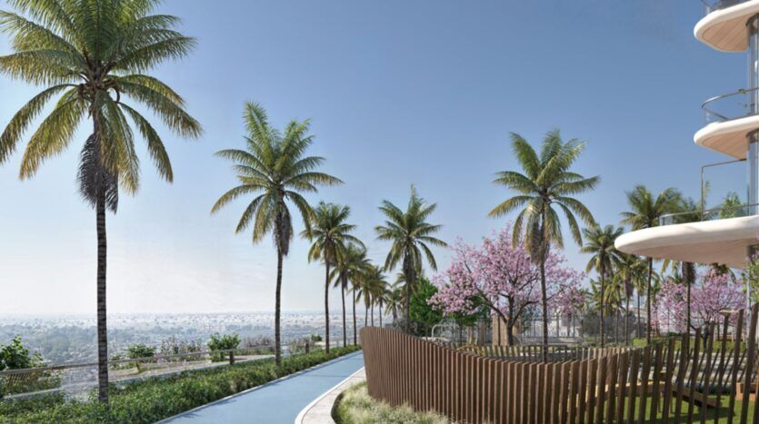 Un sentier sinueux, bordé de palmiers et d'arbres en fleurs aux fleurs roses, longe les Mövenpick Residences, un immeuble moderne doté de balcons, d'où l'on aperçoit au loin la ville sous un ciel bleu limpide.