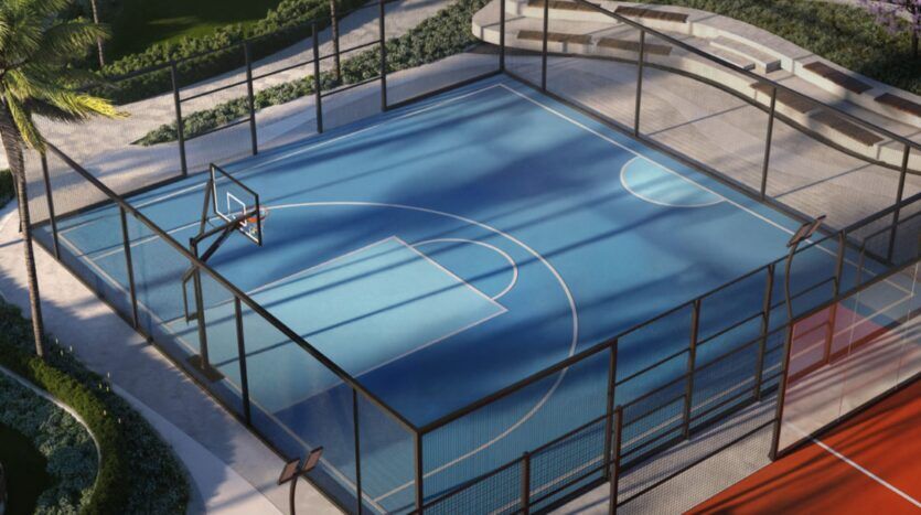 Un terrain de basket en plein air bleu, situé au sein des Mövenpick Residences, est entouré d'une haute clôture noire et d'une végétation luxuriante. La lumière du soleil et les ombres se succèdent sur le terrain désert, tandis que l'on aperçoit à proximité des bancs et des espaces verts soigneusement entretenus.