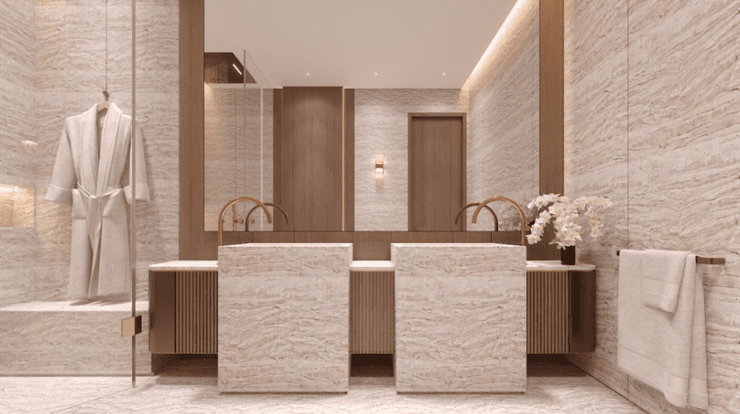 Une salle de bain moderne avec murs et sol en marbre beige. Il comprend un meuble-lavabo à double lavabo avec des accents en bois, des miroirs circulaires et un vase de fleurs blanches. Un peignoir blanc est suspendu dans la douche vitrée. Une serviette est soigneusement accrochée sur un porte-serviettes à droite.
