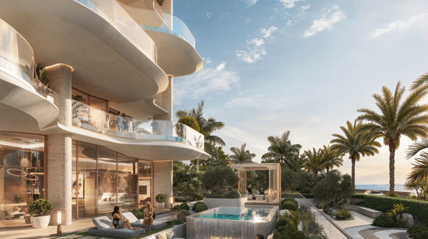 Le luxueux complexe d'appartements en bord de mer Vitalia, situé sur Palm Jumeirah, présente une architecture moderne avec des balcons incurvés. Les résidents se détendent au bord de la piscine et du coin salon, entourés de palmiers et d'une végétation luxuriante, tandis que l'océan offre une toile de fond époustouflante sous un ciel partiellement nuageux.