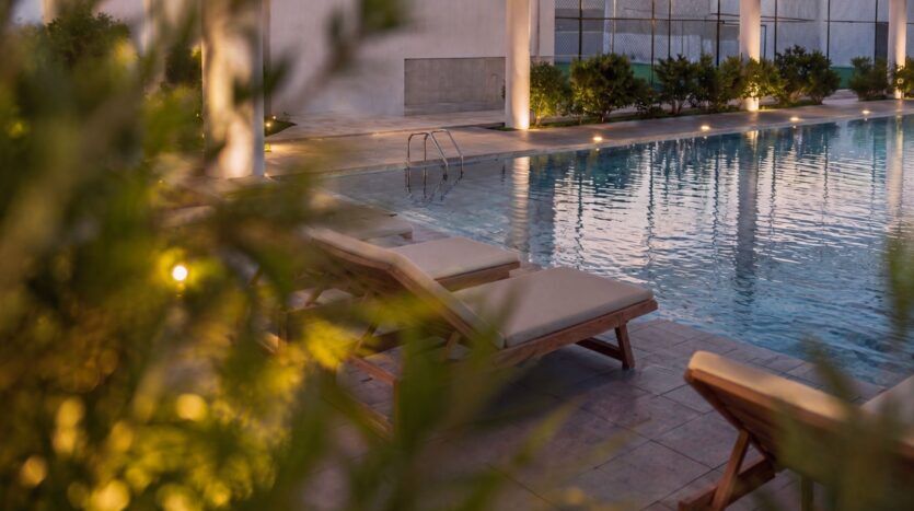 La piscine extérieure paisible du Beverly Crown by HMB est agrémentée de chaises longues rembourrées sur la terrasse, au milieu d'une végétation luxuriante et sous un éclairage tamisé en soirée.