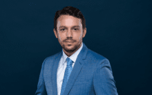 A professional photo of Guillaume Giroux with short brown hair and a light three-day beard, wearing a blue suit and patterned blue tie, against a dark blue background.