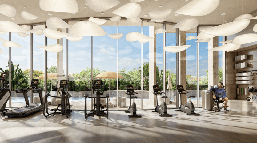 A modern gym in a Dubai villa with large windows and cloud-shaped lights fixed to the ceiling. Exercise equipment is neatly arranged and a man sits checking his phone with plants in the background.