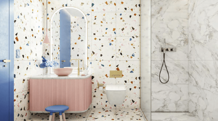 A bright, modern bathroom with terrazzo-style tiled walls, marble floor and pink basin with round mirror. Includes white toilet, glass shower and decorative accents like a blue stool and