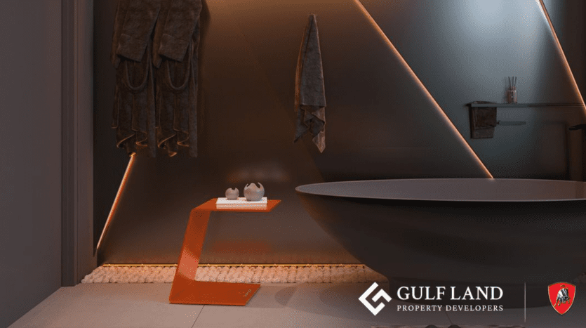 A modern bathroom with dark walls and elegant lighting. A round bathtub rests on a white pebble base next to an angular orange side table and three dark towels hung on the wall. The "Gulf Land Property Developers" logo, associated with Tonino Lamborghini Residences Dubai, is visible on the lower right.
