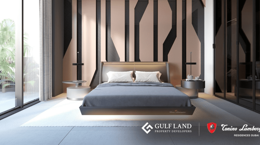 Modern bedroom with clean design. The room features a king-size bed with a gray headboard against a geometrically patterned wall. Bedside tables with lamps are on either side. Floor-to-ceiling windows let in natural light, and the Gulf Land and Tonino Lamborghini Residences Dubai logos are visible.
