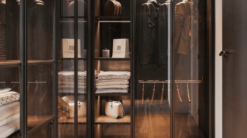 A modern closet with glass doors reveals neatly folded clothes, handbags and hats. Wooden shelves and hangers hold a variety of garments, including folded shirts, a beige handbag and hanging coats. The overall aesthetic of Tonino Lamborghini Residences Dubai is minimalist and organized.