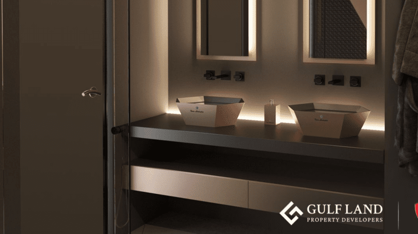 A modern bathroom with soft ambient lighting features an elegant black countertop with two square-shaped sinks. There are two mirrors above the sinks and towel hooks on the right-hand wall. The "Gulf Land Property Developers" logo is in the lower right-hand corner, alluding to Tonino Lamborghini Residences Dubai.