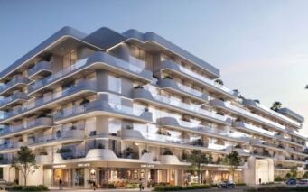 Un immeuble moderne de plusieurs étages - Oxford Cove by Iman - est doté de balcons blancs et épurés, de garde-corps en verre et de palmiers sur les terrasses. Des boutiques et des cafés bordent le rez-de-chaussée, avec des voitures garées et des gens qui se promènent à proximité au coucher du soleil.