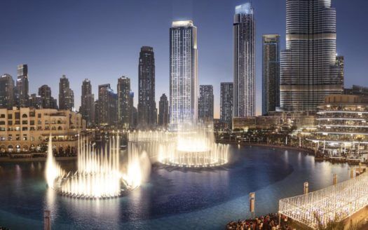 Vista panorámica de un paisaje urbano al atardecer con rascacielos emblemáticos que dominan un gran espectáculo de fuentes, luces que se reflejan en el agua y multitudes que observan desde el paseo marítimo.